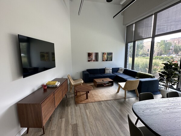 A modern living room with a sectional sofa, a flat-screen TV mounted on the wall, a wooden coffee table, and a dining table with chairs.