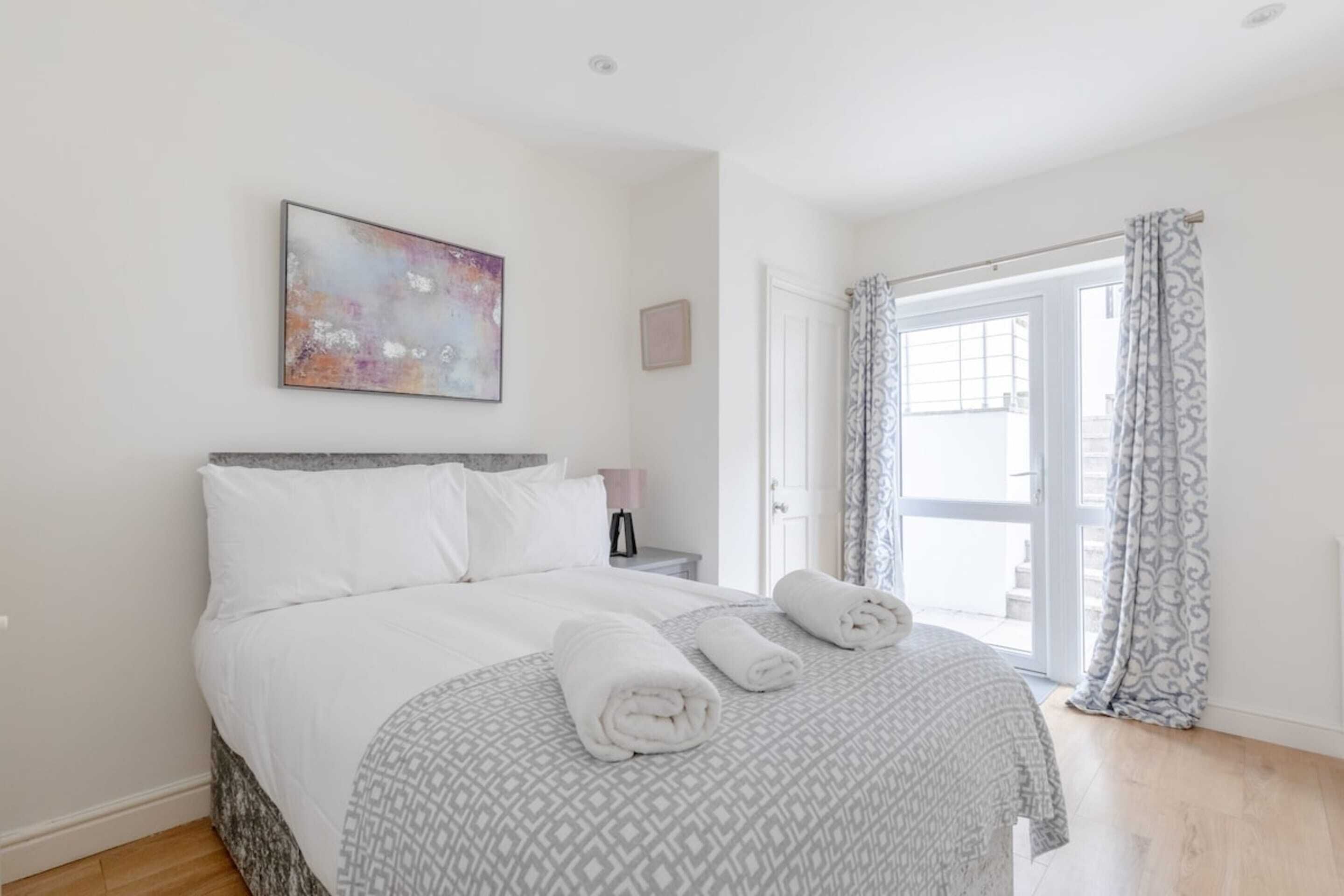 Un lit bien fait, recouvert d’une couvre-lit à motifs, une table de chevet avec une lampe et une grande fenêtre avec des rideaux.