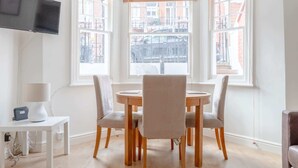 A dining area with a round table and four chairs, a TV mounted on the wall, and large windows with blinds. - Beautifully Presented 2BD Flat W/patio- Kensington (London)