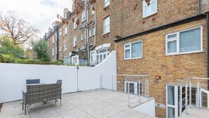 A residential building with a tiled patio, a wicker bench, and a white wall. - Beautifully Presented 2BD Flat W/patio- Kensington (London)