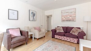 A living room with a brown leather sofa, a white armchair, and a purple sofa with patterned cushions. There are framed artworks on the walls and a white door in the background. - Beautifully Presented 2BD Flat W/patio- Kensington (London)