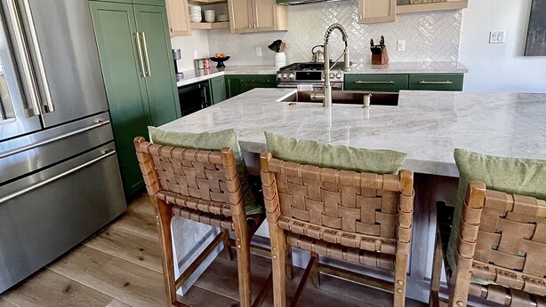 Une cuisine avec des meubles de couleur verte, un réfrigérateur en acier inoxydable, un îlot en marbre avec quatre chaises en osier et des luminaires suspendus.