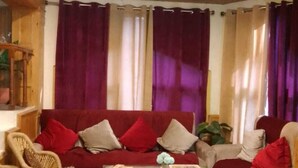 A cozy living room with a red sofa, two armchairs, a wooden coffee table, and a ceiling with a wooden panel design. - Hotel Anupam  (Bhuntar)