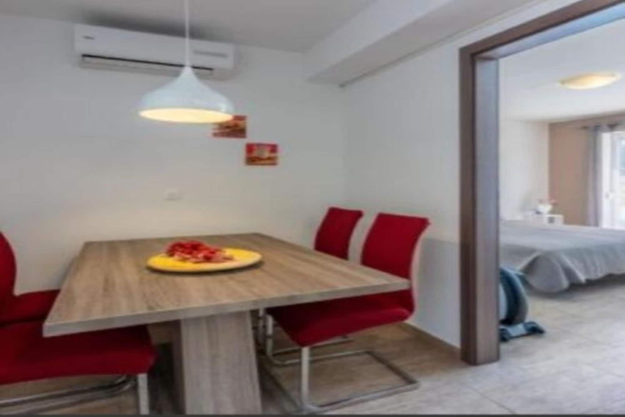 A compact hotel room with a dining area featuring a wooden table and red chairs, a bed with a grey bedspread, and a visible air conditioning unit.