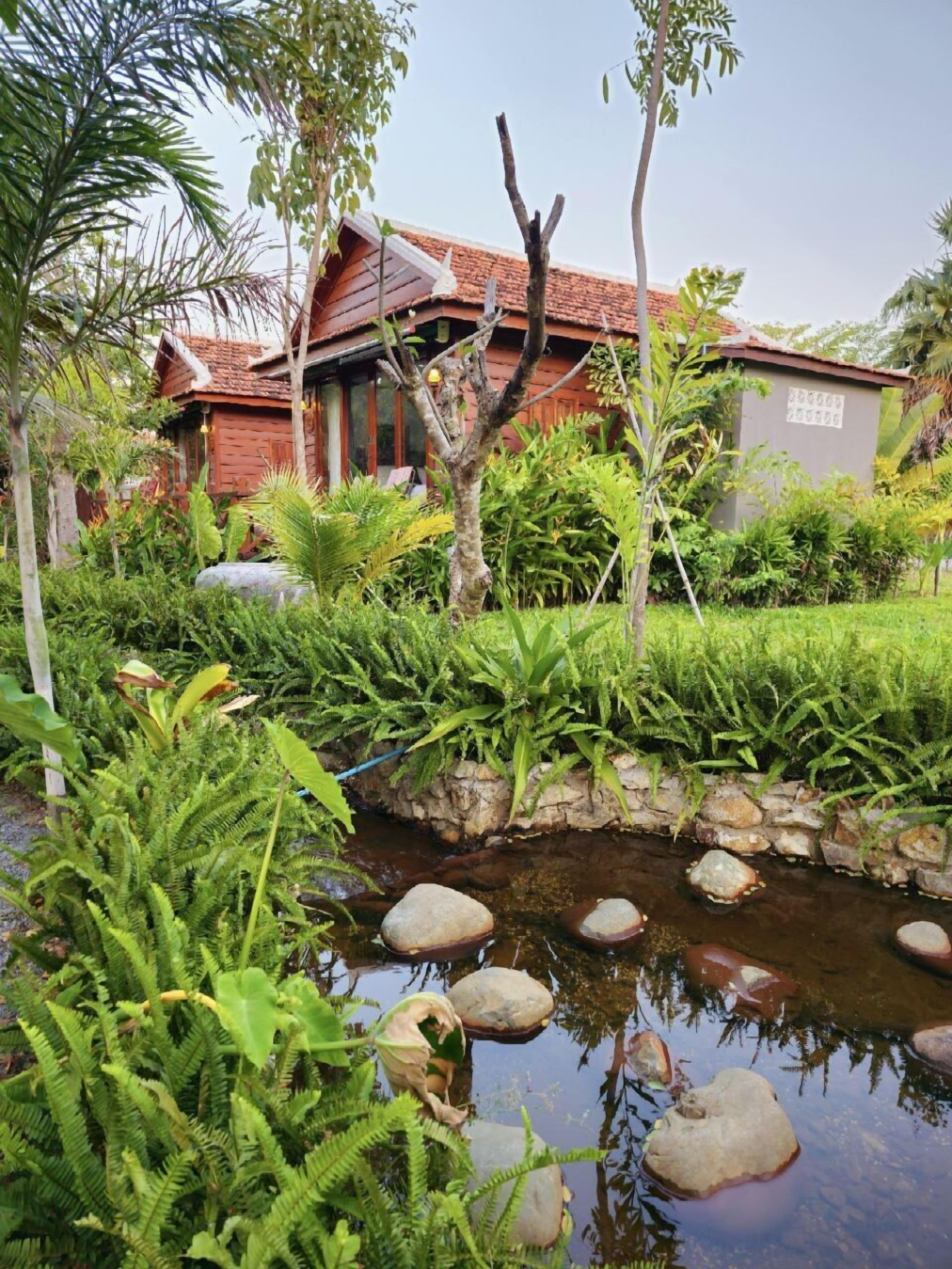 A traditional house with a tiled roof, surrounded by lush greenery and a small stream with rocks.