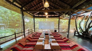 Un salón de conferencias con una larga mesa, cojines estampados en rojo y beige, y una pantalla de proyector al frente.