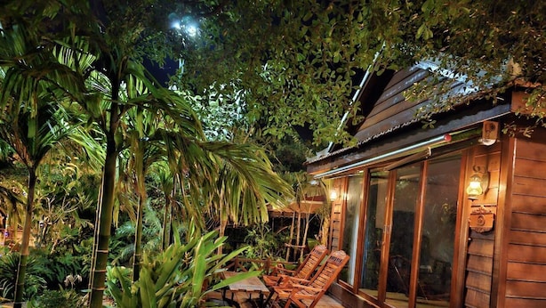 Una terraza de madera con sillones de descanso y una sala con paredes de vidrio, rodeada de exuberante vegetación tropical.