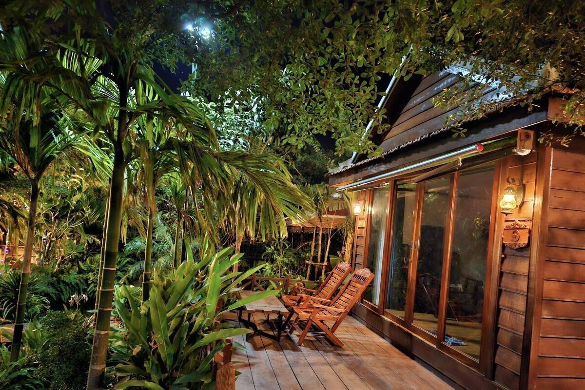 Une terrasse en bois avec des chaises longues et une pièce fermée par des baies vitrées, le tout entouré d’une végétation tropicale luxuriante.