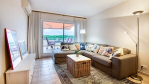 Una sala de estar moderna con un sofá, una mesa de centro y una lámpara de pie. Hay un ventanal con cortinas y vistas al exterior con vegetación.
