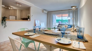 Un comedor con una mesa de madera preparada para cuatro, vista al balcón y una cocina al fondo.