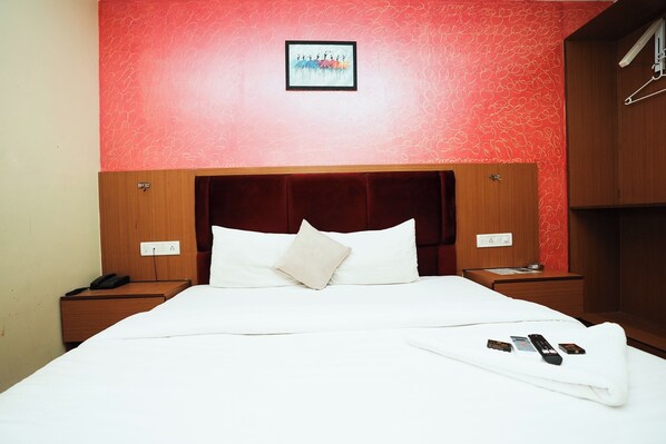 A hotel room with a red wall, a framed picture, a wooden headboard, a white bed, two bedside tables, and a blanket with remote controls. - Hotel Twigo Inn (Mumbai)