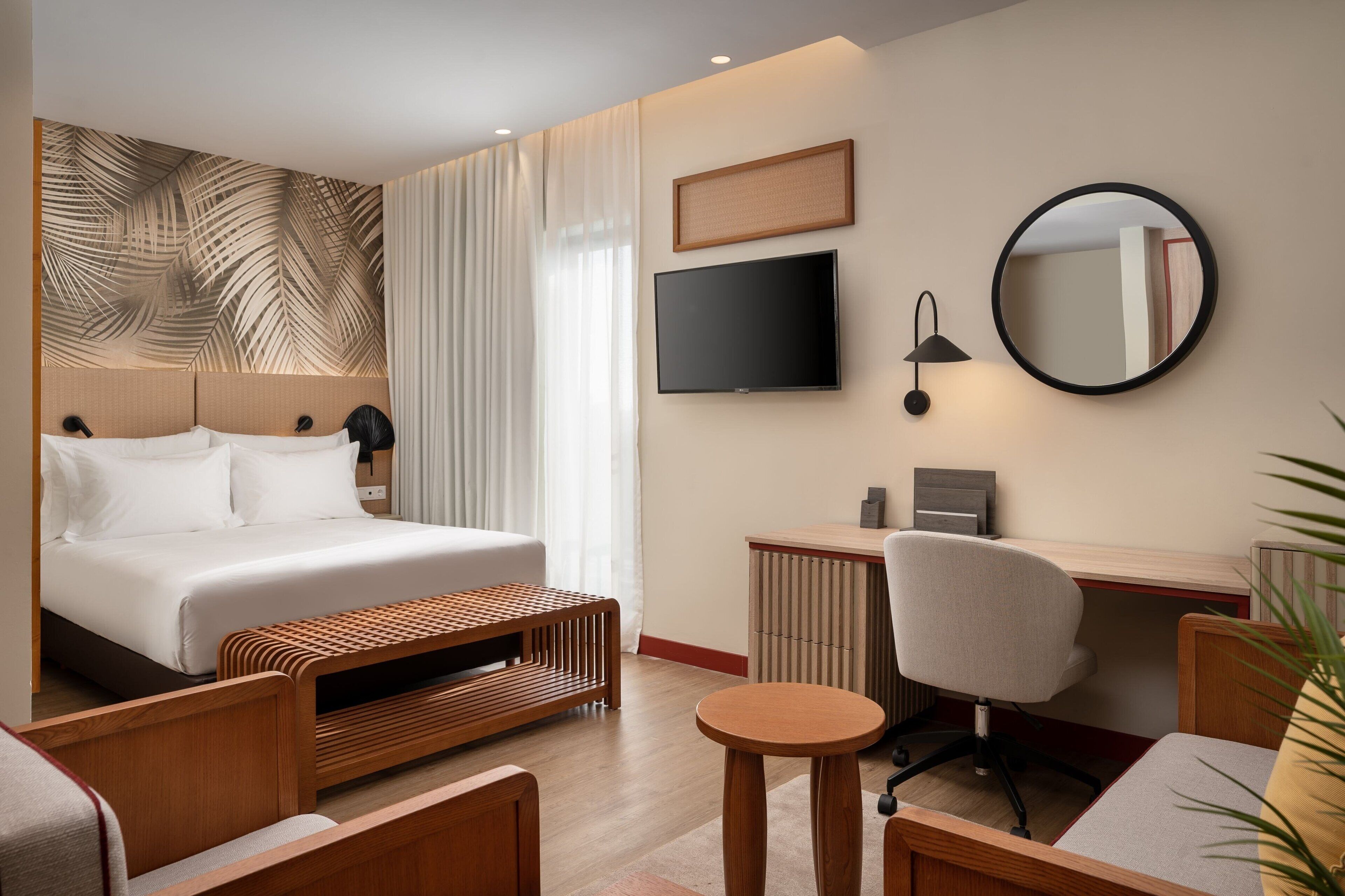 Una habitación de hotel moderna con una cama grande, un escritorio, una silla, un espejo redondo y un televisor.