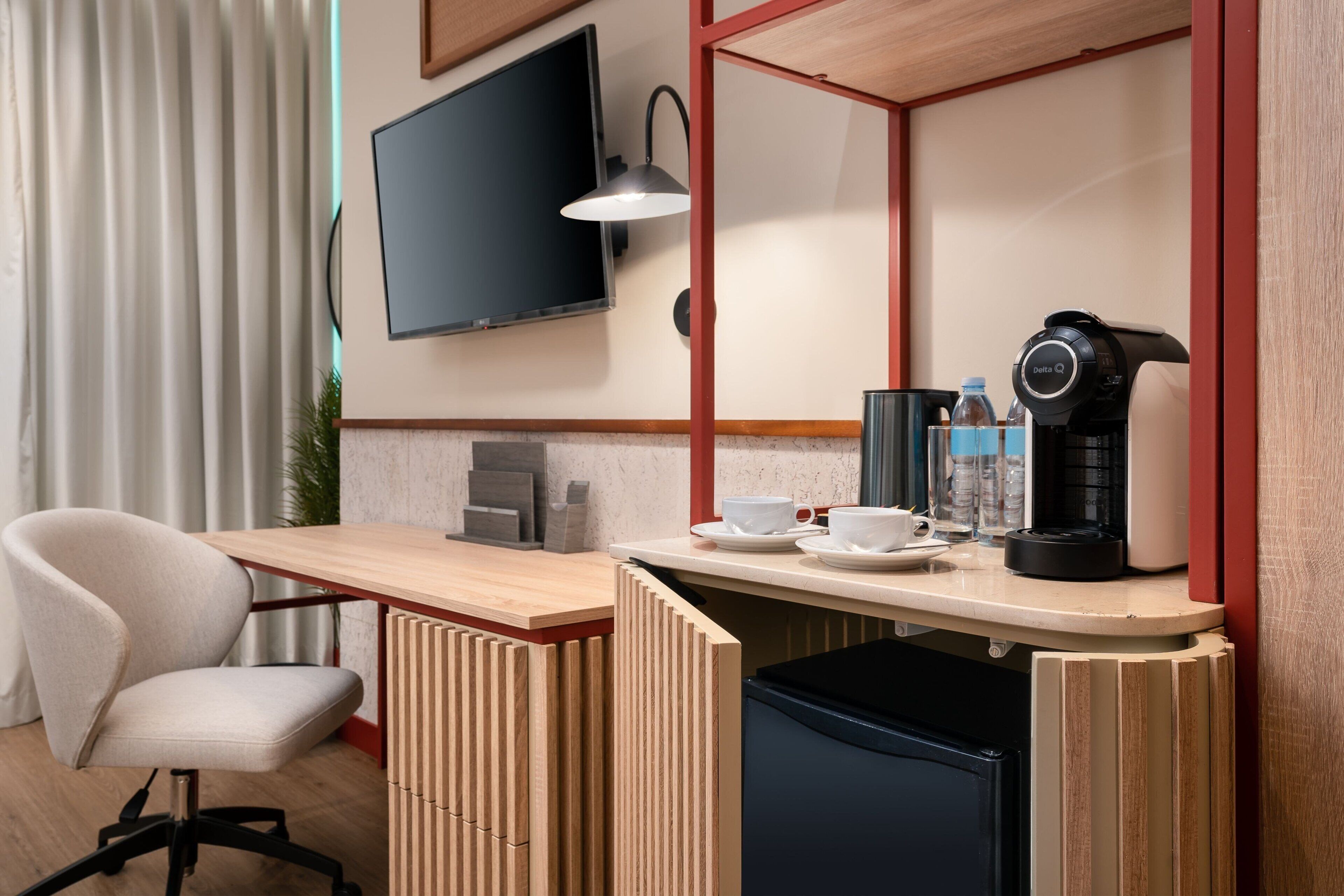 Ein kleines Hotelzimmer mit Schreibtisch aus Holz, einem Sessel, einem Fernseher, einer Kaffeemaschine und einem kleinen Kühlschrank.