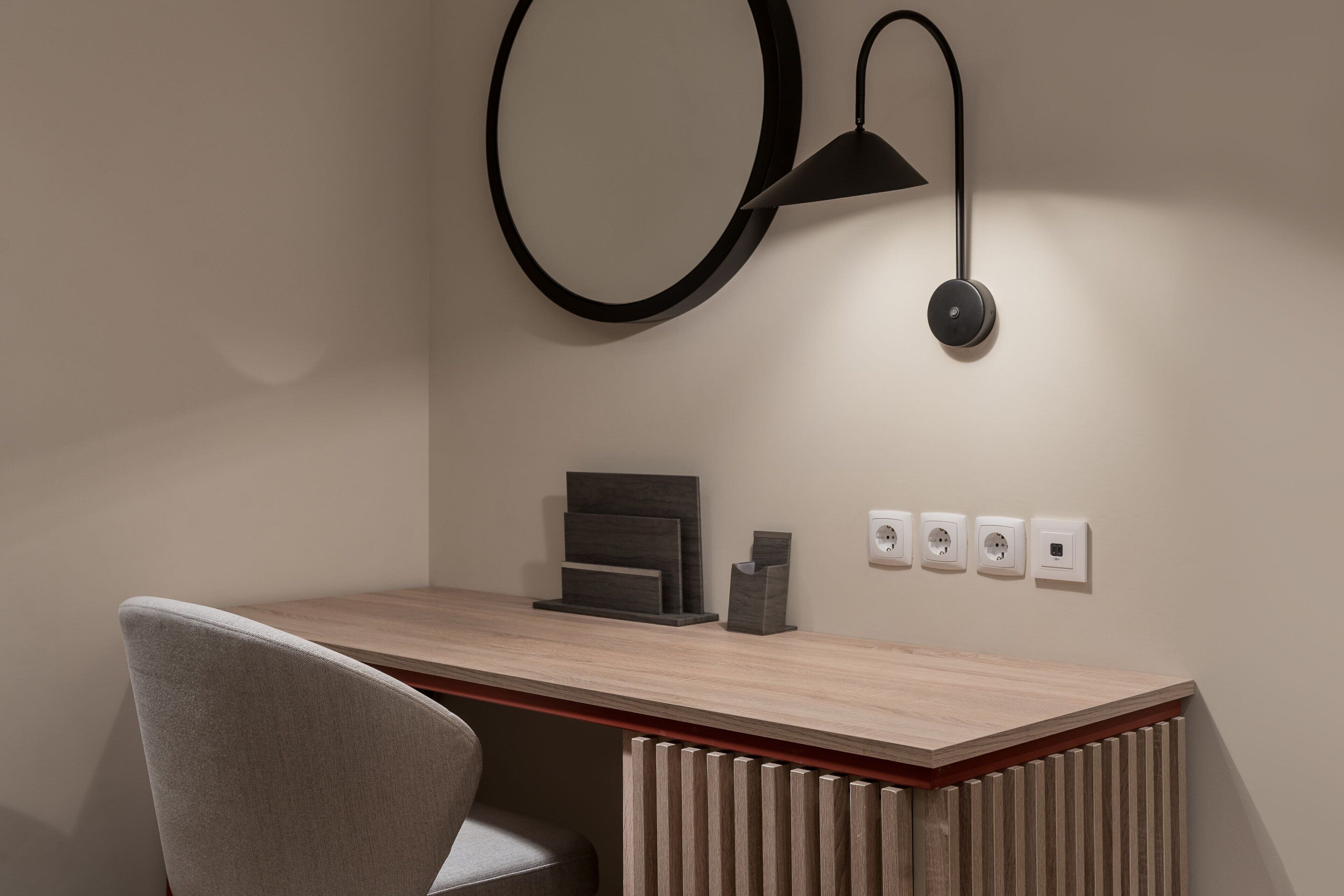 A modern desk with a light wood finish, a grey upholstered chair, a black wall-mounted lamp, and a minimalist decor piece.