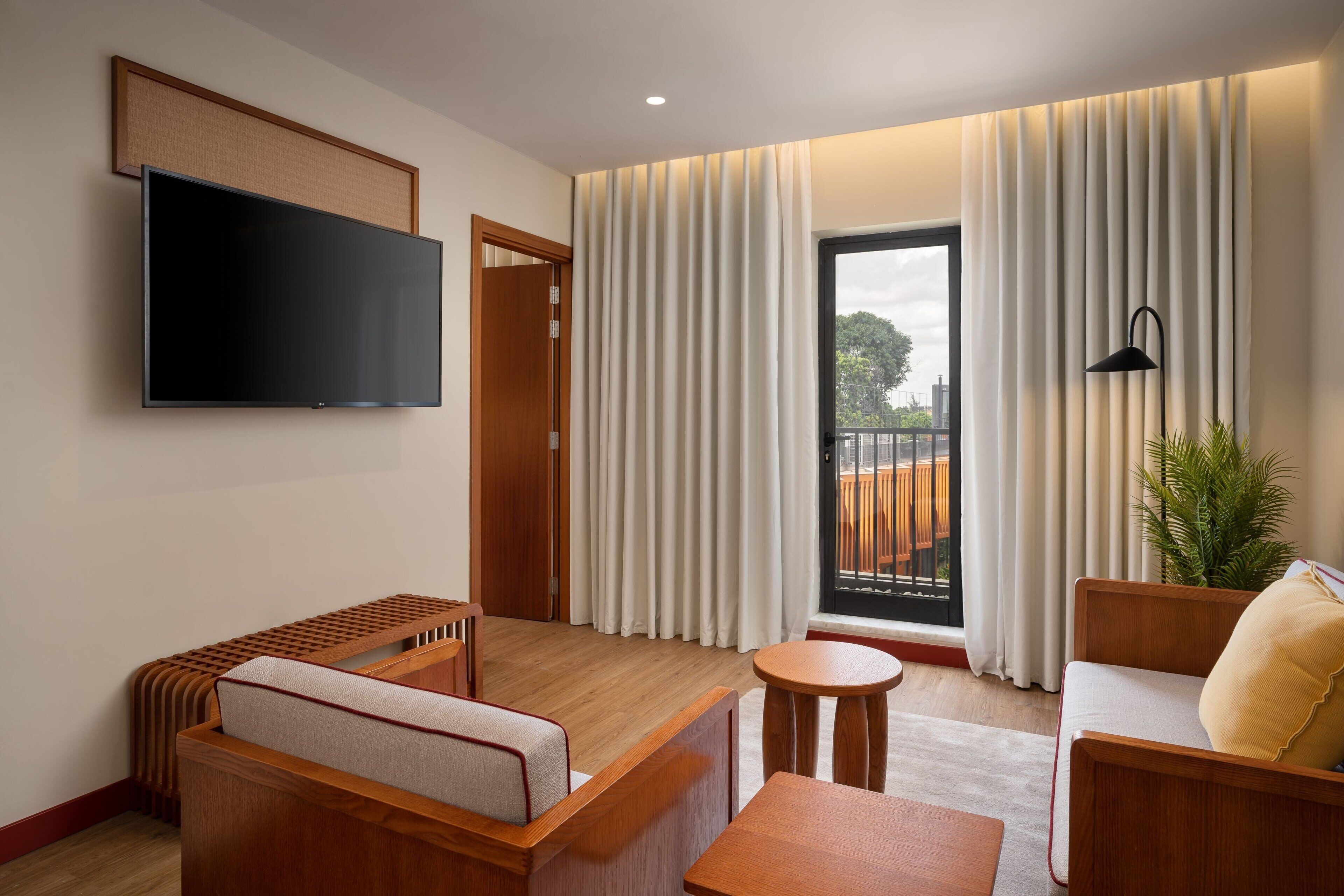 A modern hotel room with a flat-screen TV, a sofa, a small table, and a balcony with curtains.