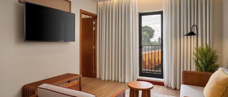 A modern hotel room with a flat-screen TV, a sofa, a small table, and a balcony with curtains.