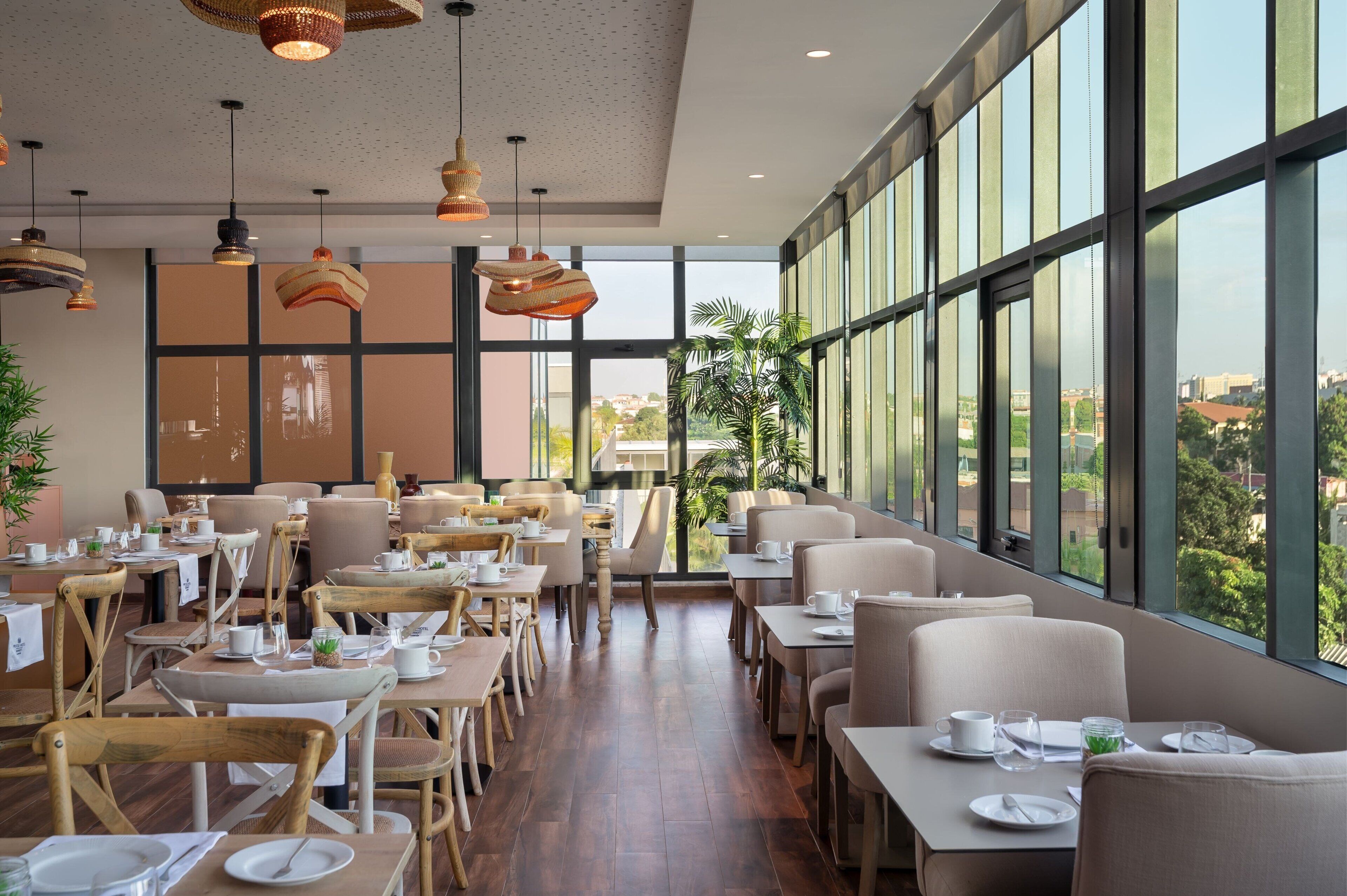 Un'ampia sala da pranzo con soffitti alti, ampie finestre e lampade a sospensione moderne. I tavoli sono disposti ordinatamente con sedie, e si può ammirare una vista che include piante e palazzi.