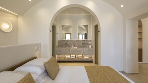 A modern bedroom with a large bed, a bathroom with a marble countertop, and an arched doorway. - Scenic Pool Villa with Sunset & Sea View by Santo Collection, The Villas (Santorini)