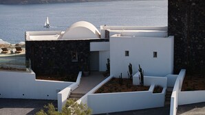 A modern building with a white facade, a stone section, and a view of the sea. - Scenic Pool Villa with Sunset & Sea View by Santo Collection, The Villas (Santorini)