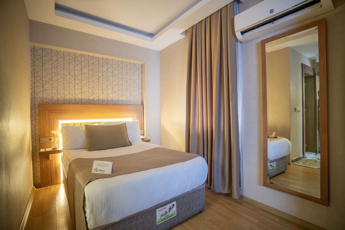 A hotel room with a bed, a nightstand, a wall-mounted air conditioner, and a large mirror.