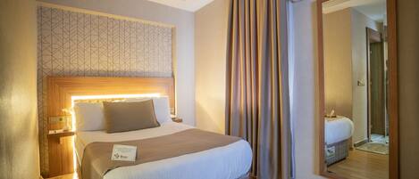 A hotel room with a bed, bedside table, lamp, curtains, and a large mirror.
