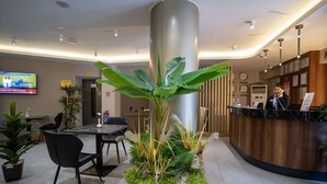 A reception area with a wooden reception desk, a large potted plant, a television mounted on the wall, and a seating area with chairs and a table. - Uğurlu Hotel (Gaziantep)