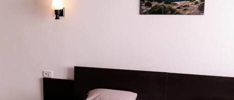 A bedroom with a bed, a nightstand, a wall-mounted lamp, and a painting of a coastal landscape.