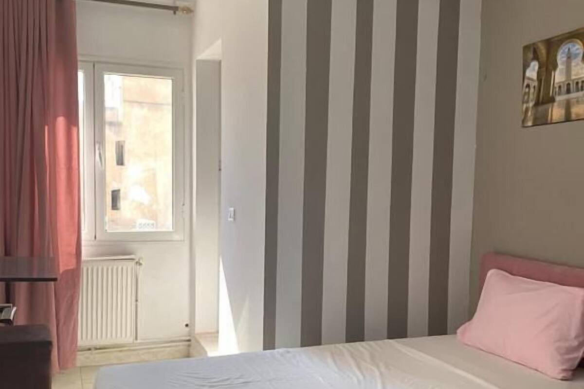 A bedroom with a bed, a radiator, a window with curtains, and a wall with vertical stripes.