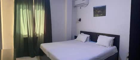 A hotel room with a bed, curtains, an air conditioning unit, and a small painting on the wall.