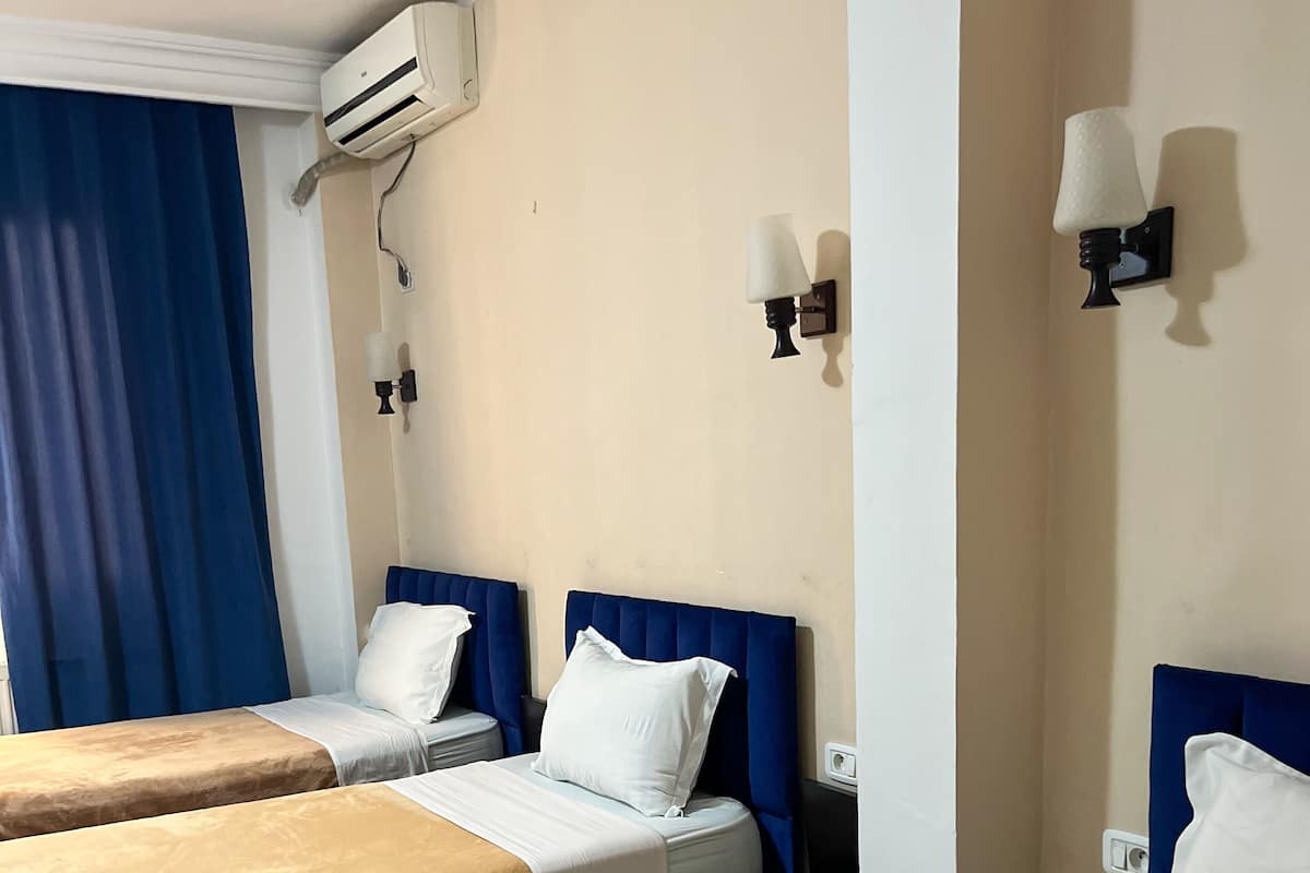 A hotel room with two beds, a blue headboard, white bedding, an air conditioning unit, and wall-mounted lamps.