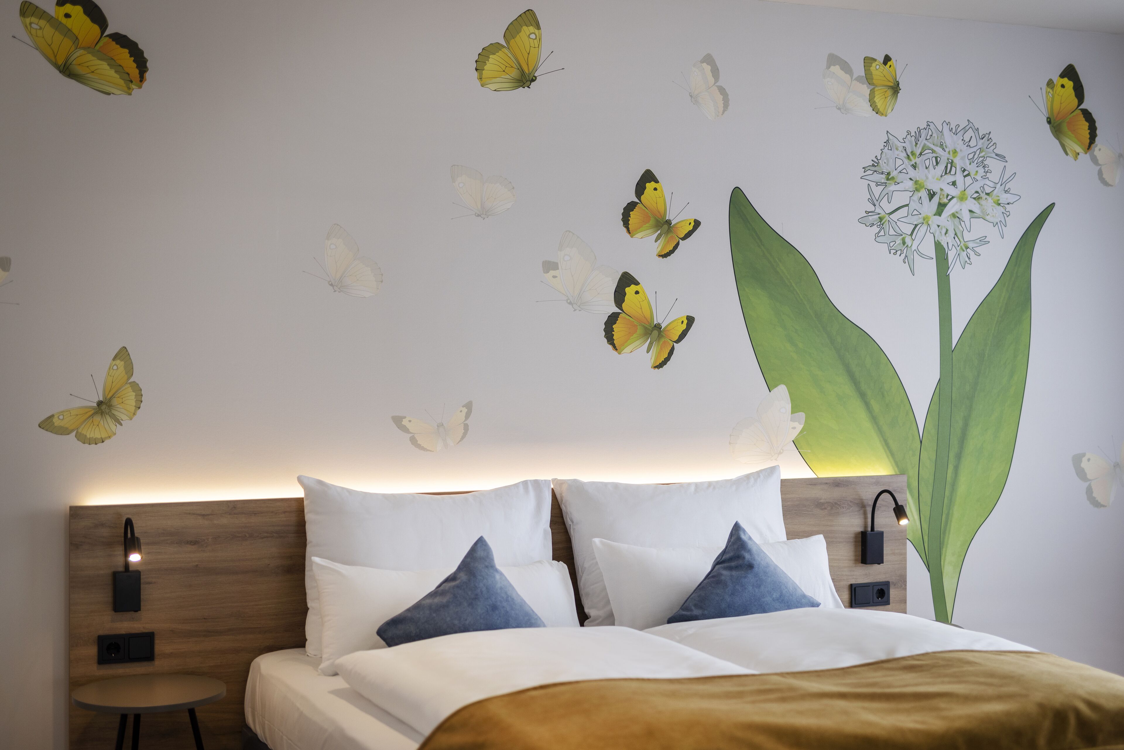 Quarto com cama, almofadas, mesa de cabeceira, luminária na parede e adesivos de borboletas.