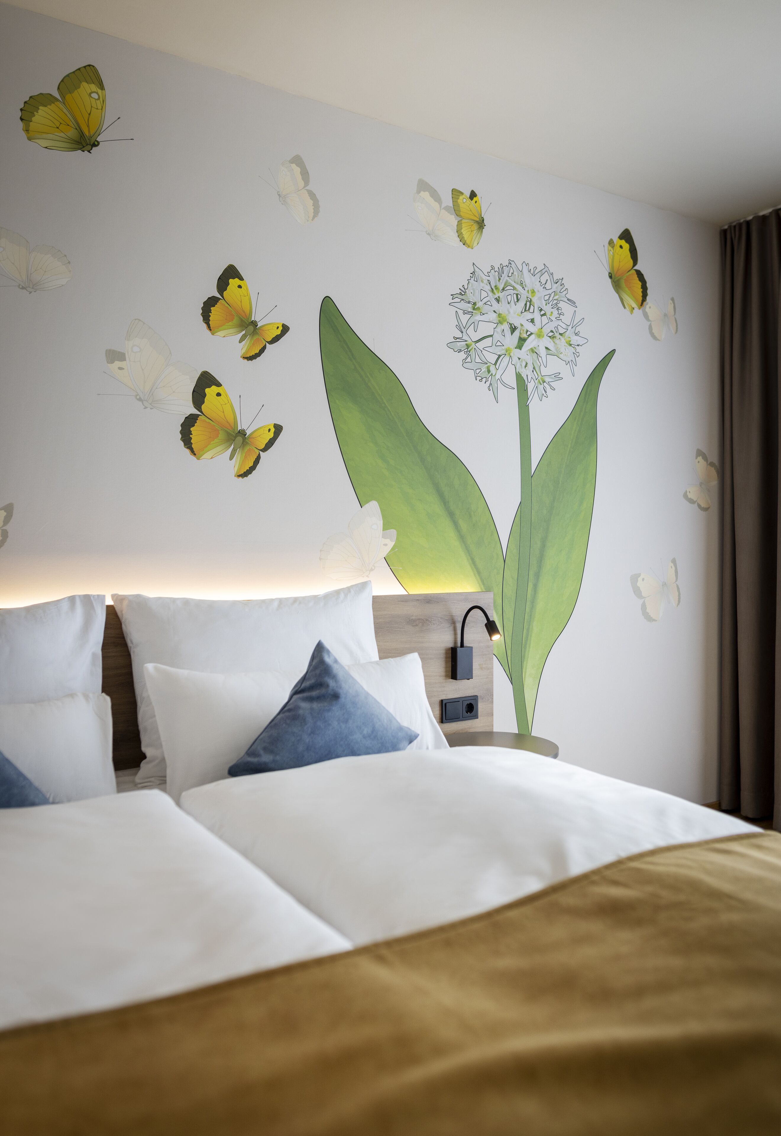 A hotel room with a bed, pillows, a nightstand, and a wall mural of butterflies and a plant.