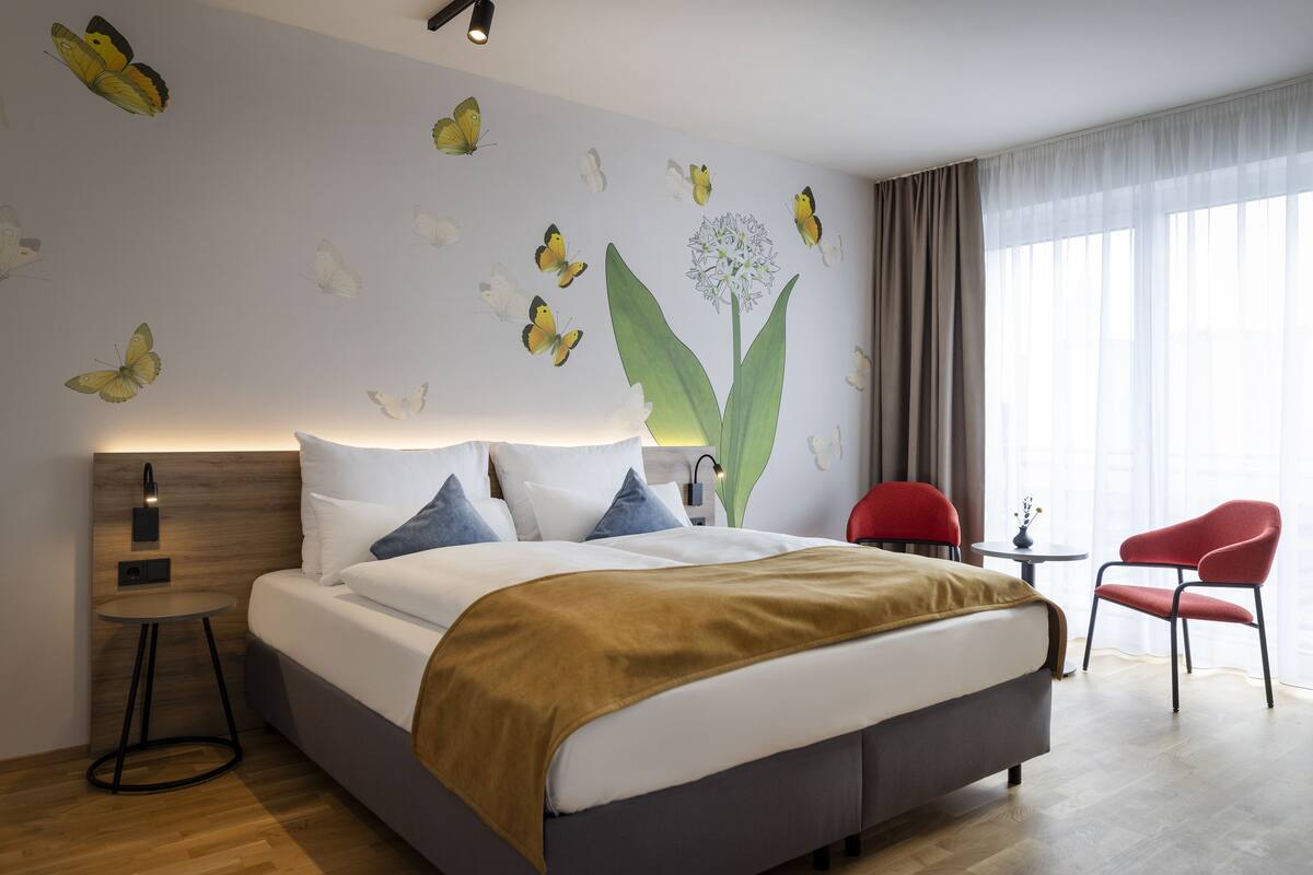 Ein modernes Hotelzimmer mit einem großen Bett, einem Nachttisch, einem roten Sessel und einem Fenster mit transparenten Vorhängen.