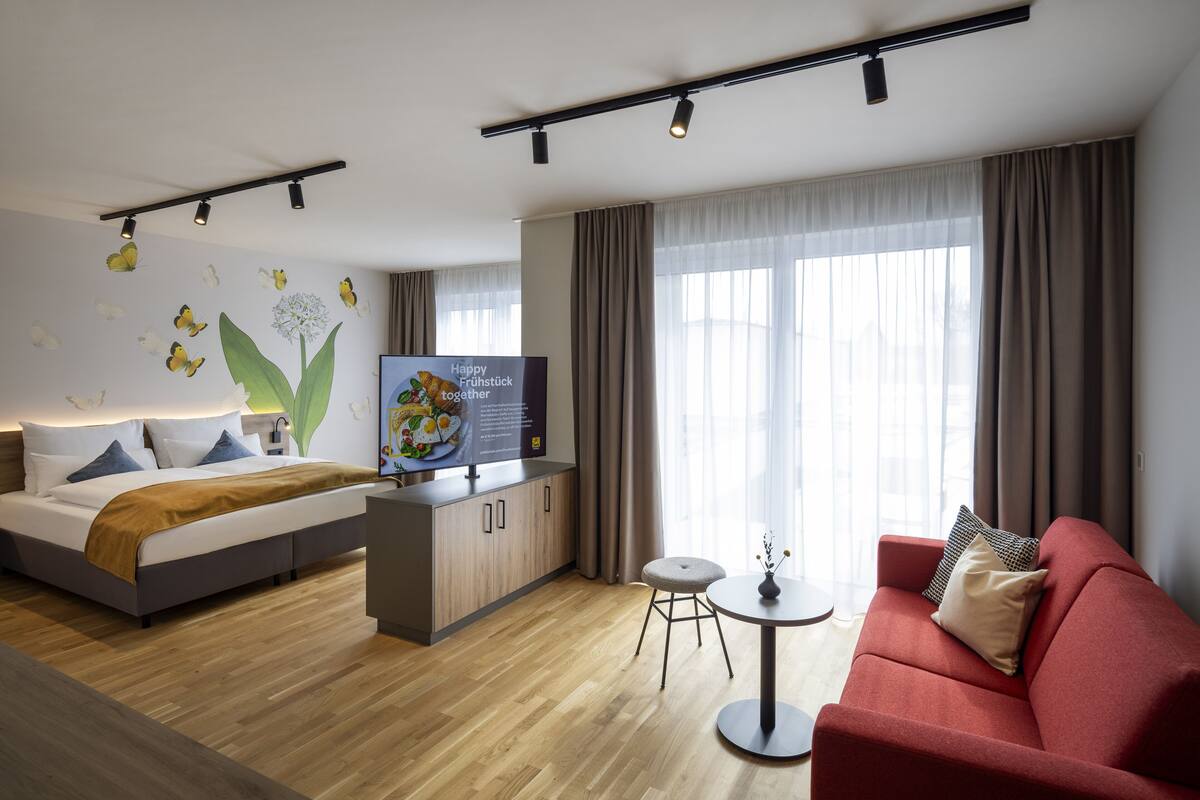 Ein modernes Hotelzimmer mit einem Bett, einem roten Sofa, einem kleinen runden Tisch, einer Küchenzeile und einem großen Fenster mit Vorhängen.