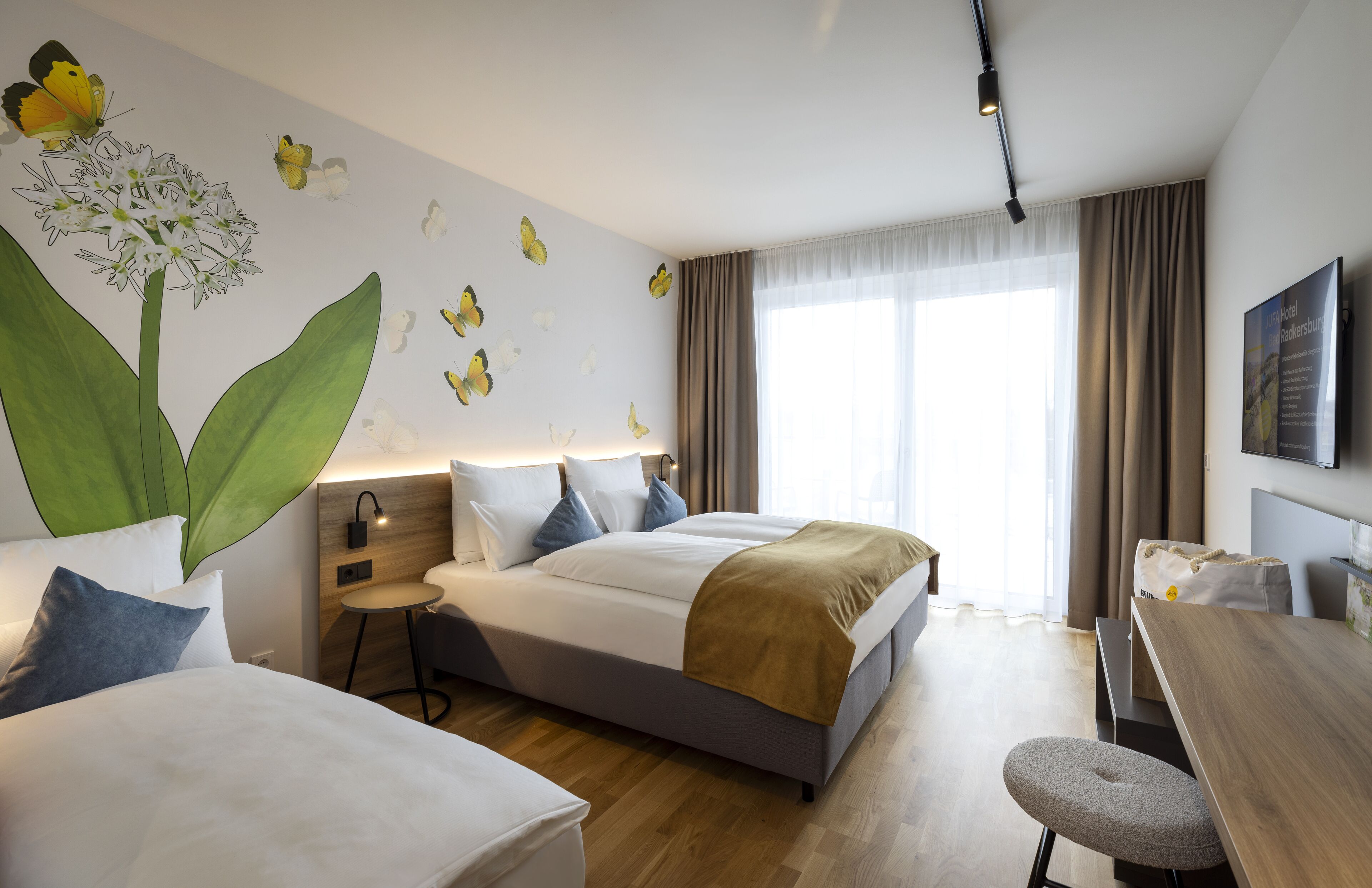A hotel room with a large bed, a smaller bed, a desk, and a chair. There is a wall mural of flowers and butterflies.