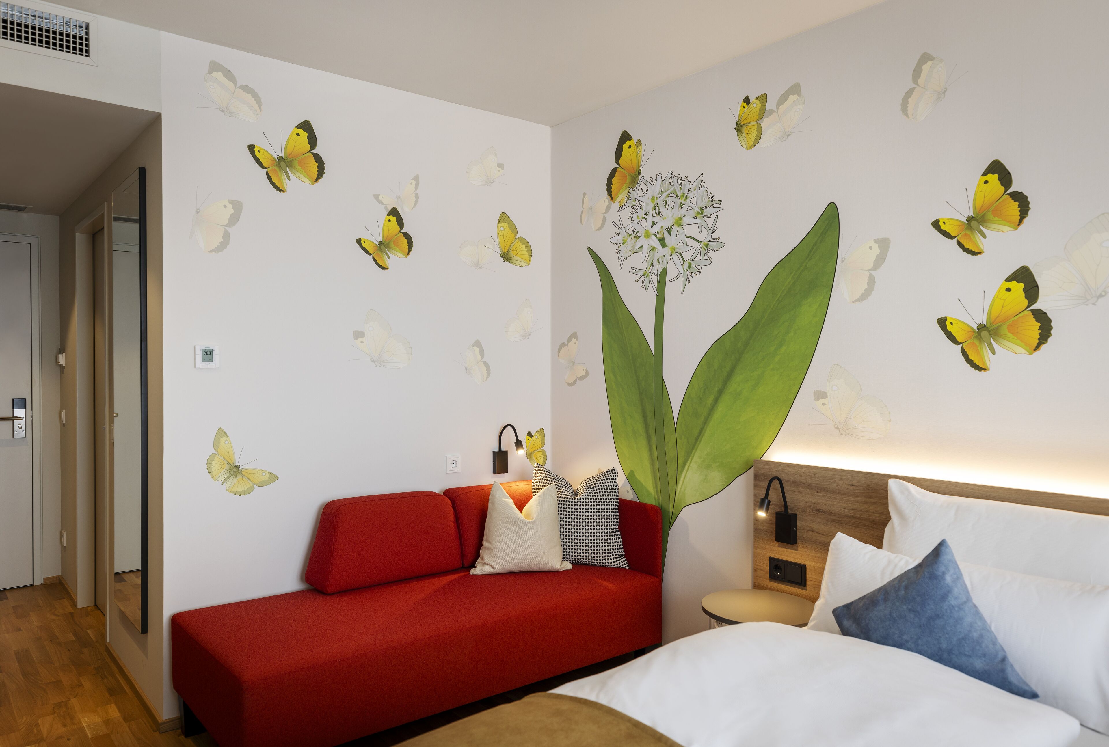 A room with a red sofa, a bed with white and blue pillows, and a wall decorated with butterfly decals and a large green plant.