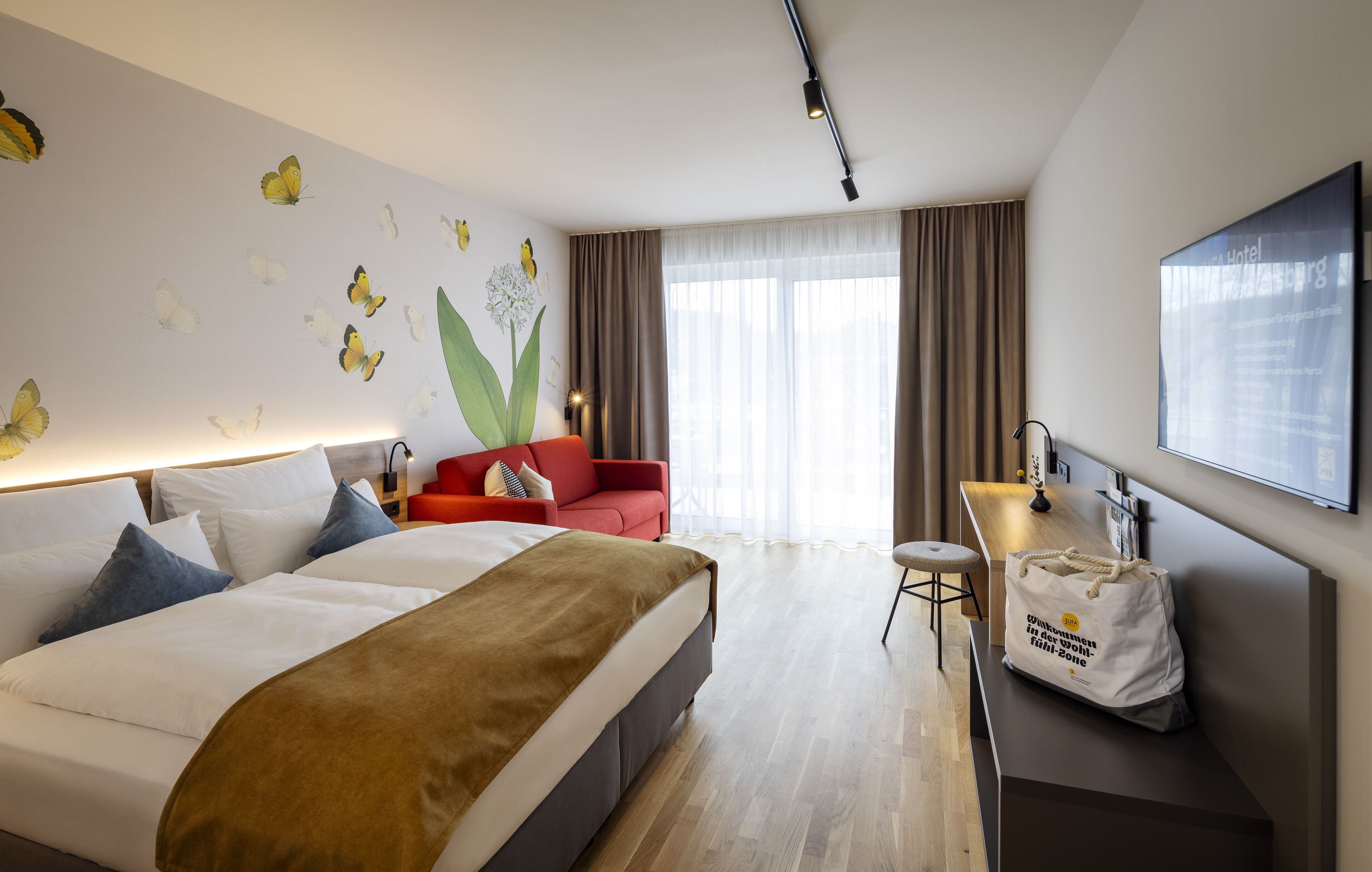 A hotel room with a bed, a red armchair, a desk with a lamp, and a large window with curtains.