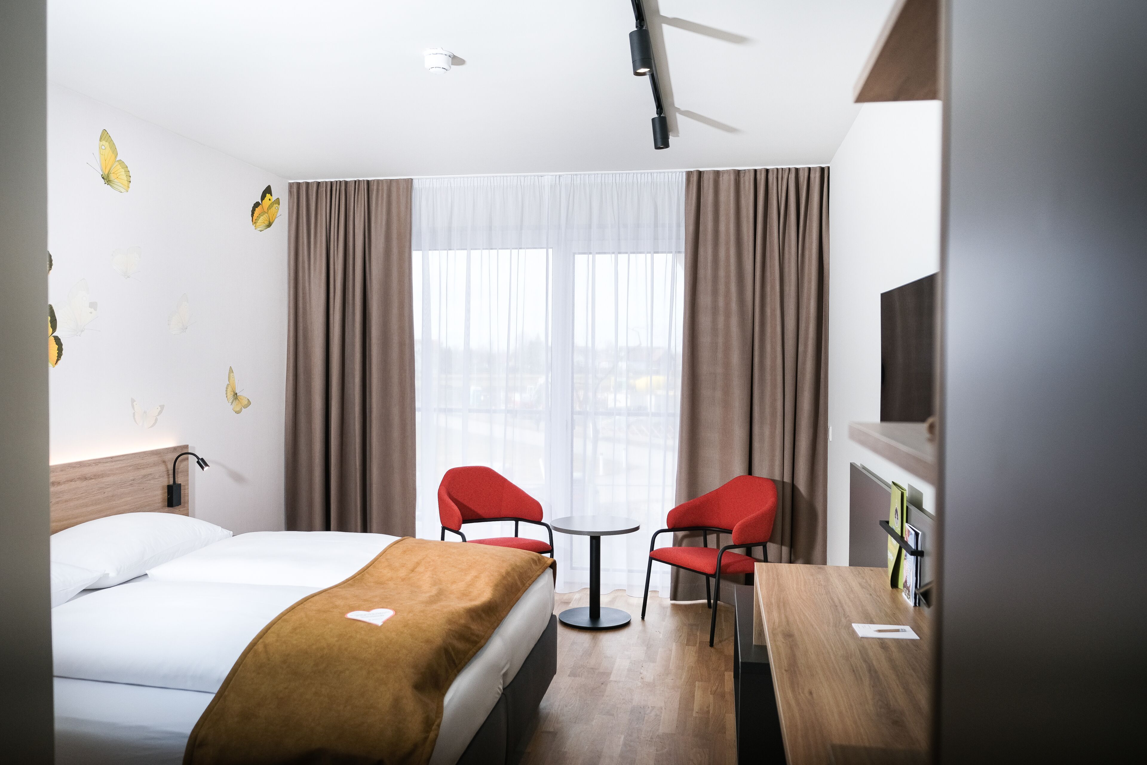A hotel room with a bed, two red chairs, a small table, a TV, and a window with curtains.