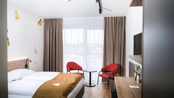 A hotel room with a bed, two red chairs, a small table, a TV, and a window with curtains.