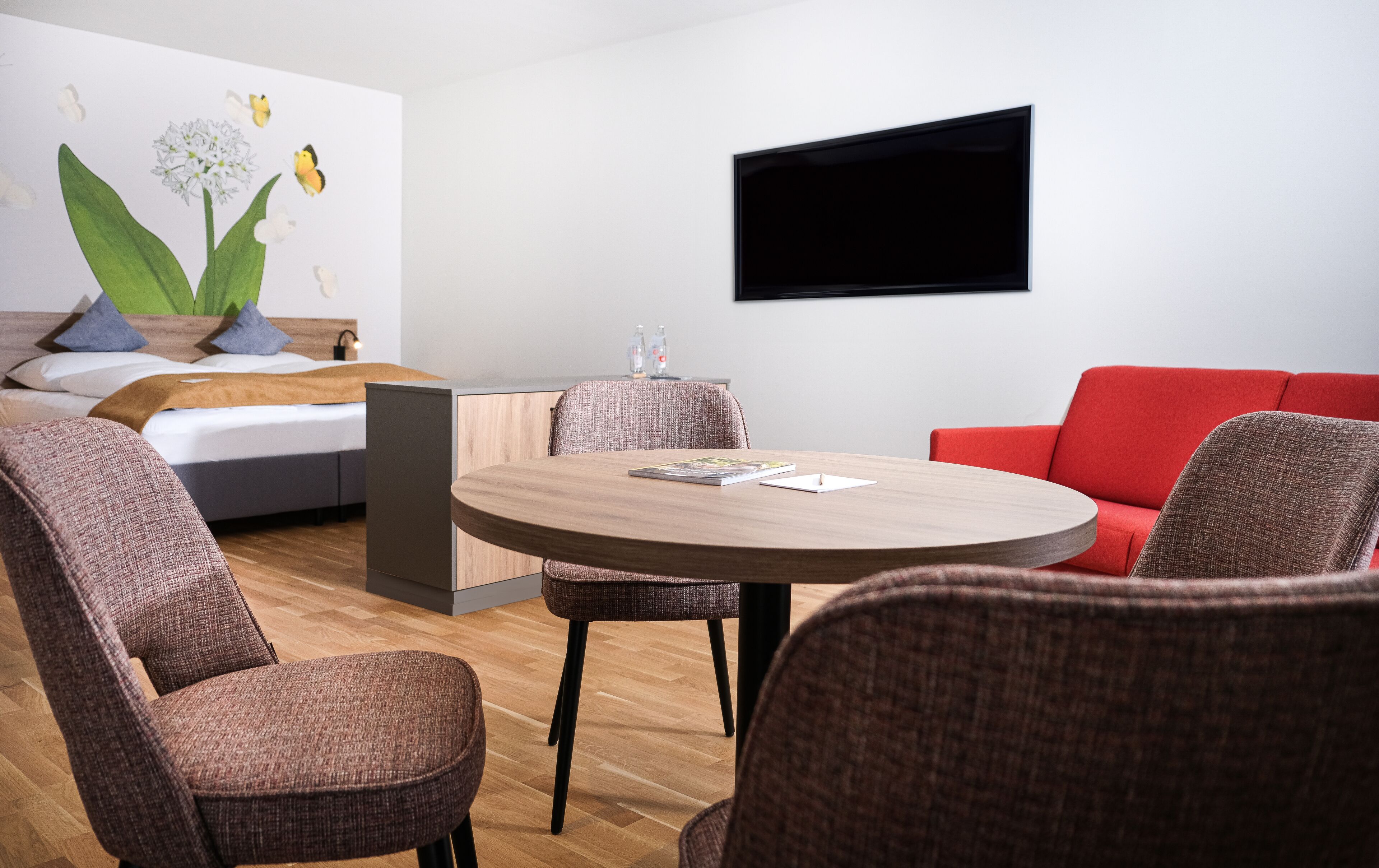 Une chambre d’hôtel moderne avec un lit, un téléviseur fixé au mur, une table ronde en bois avec des chaises et un canapé rouge.