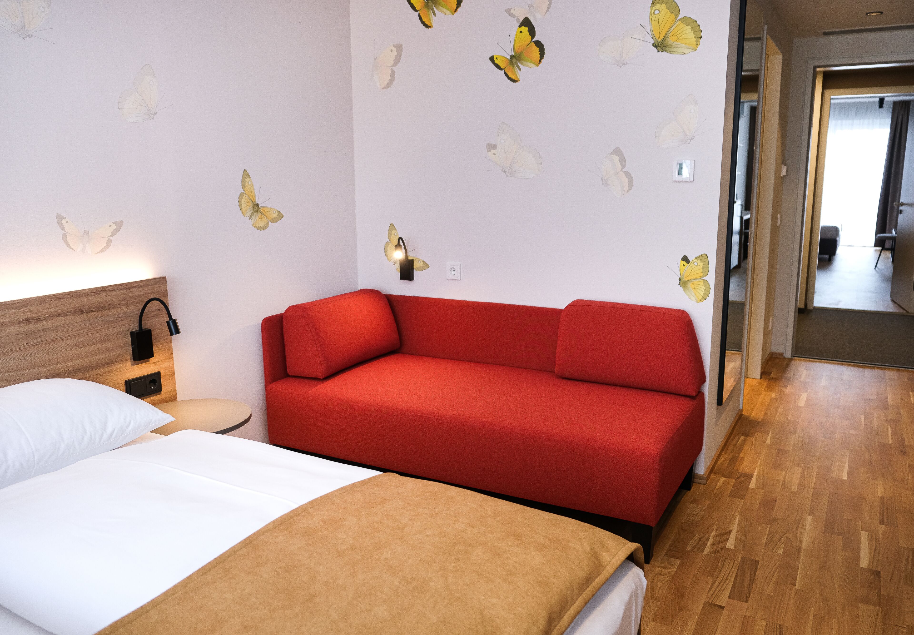 Habitación de hotel con sofá rojo, cabecera de madera, cama blanca y adhesivos de pared con mariposas.