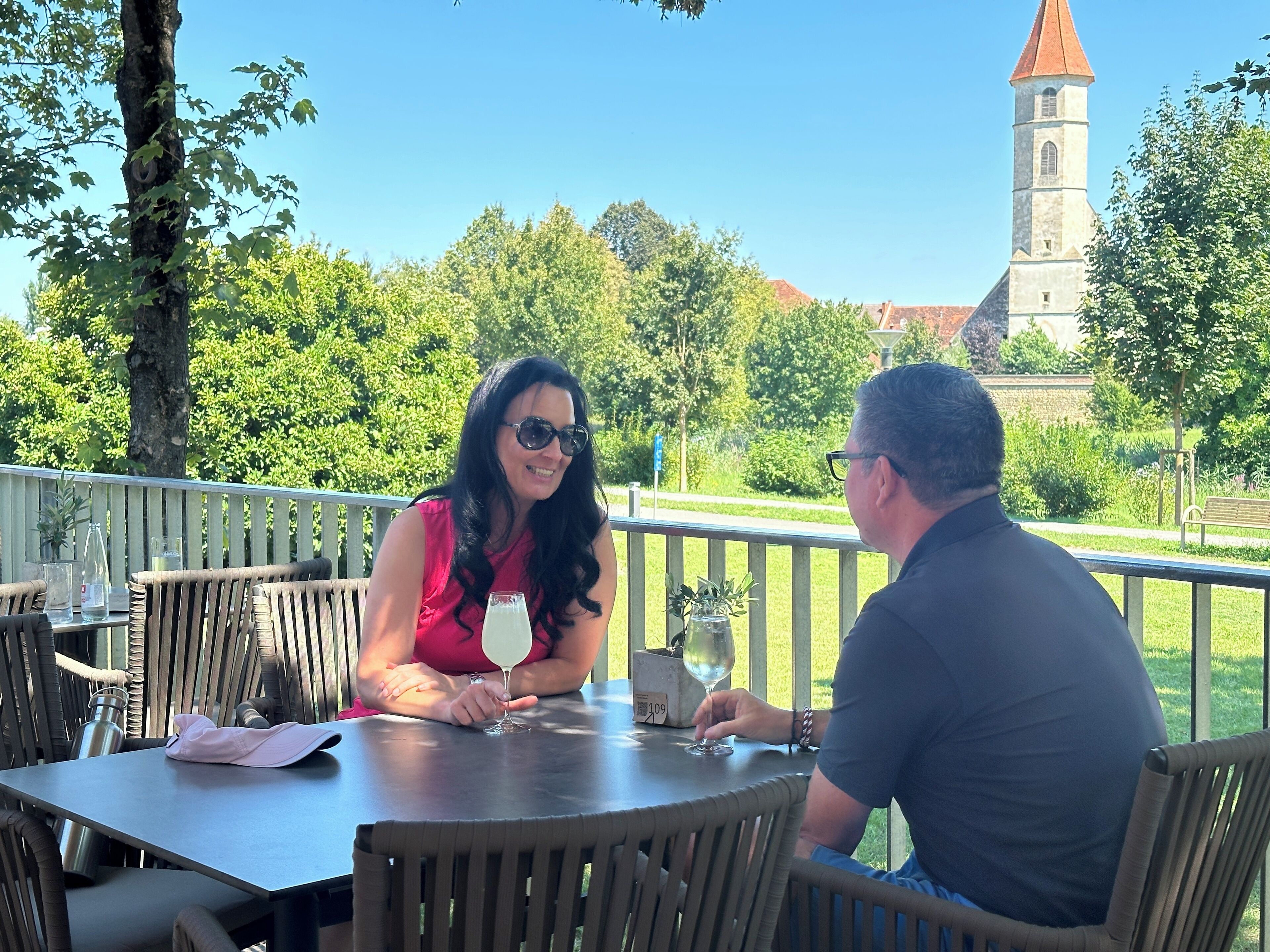 Zwei Personen sitzen an einem Tisch im Freien mit Blick auf einen Kirchturm im Hintergrund.