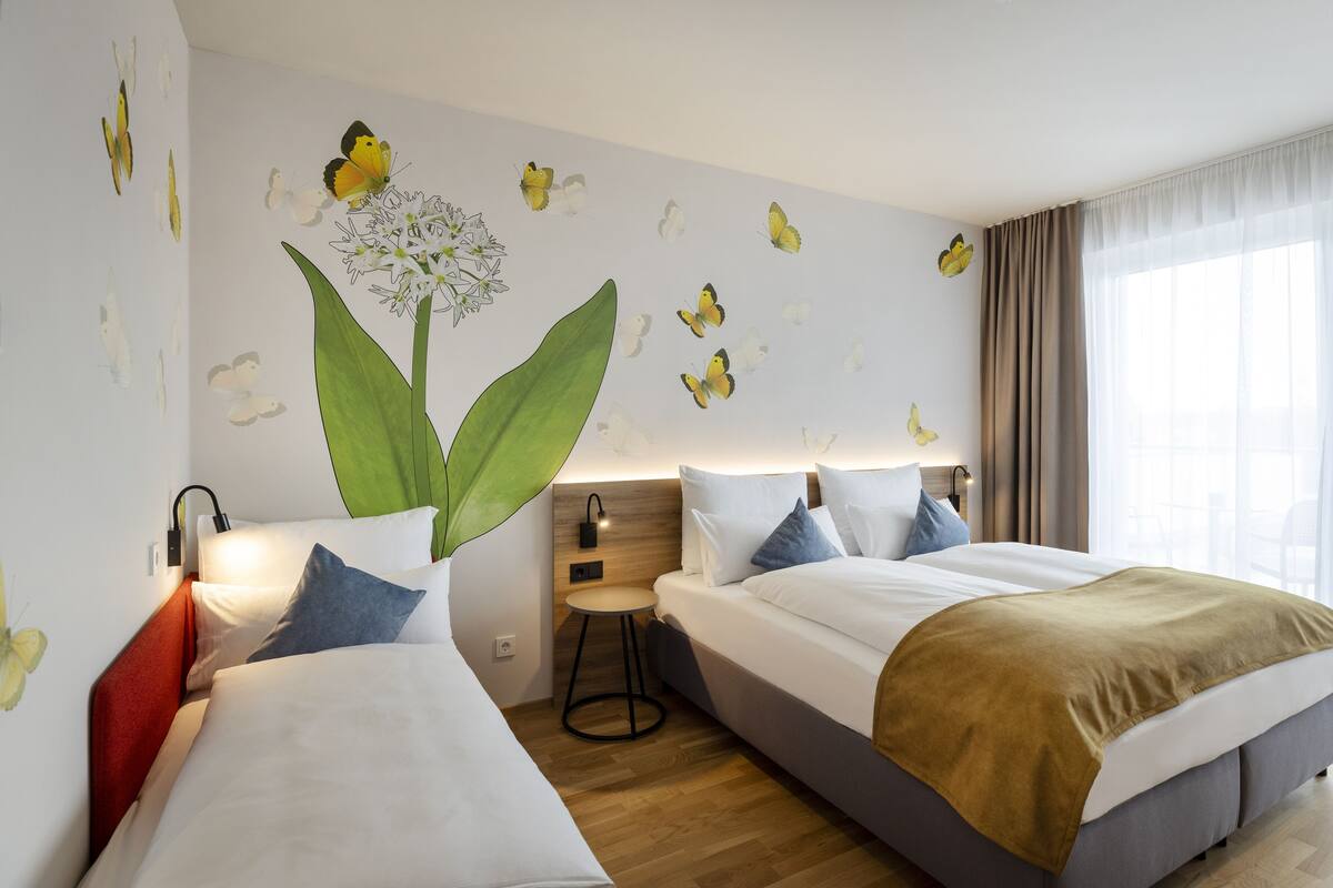Ein Hotelzimmer mit zwei Betten, einem Nachttisch, einer Lampe und einem großen Fenster mit Vorhängen.