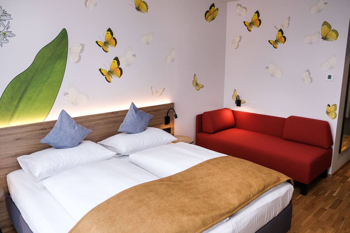 Ein Schlafzimmer mit Bett, Kissen, einem roten Sofa und Schmetterlingsstickern an der Wand.