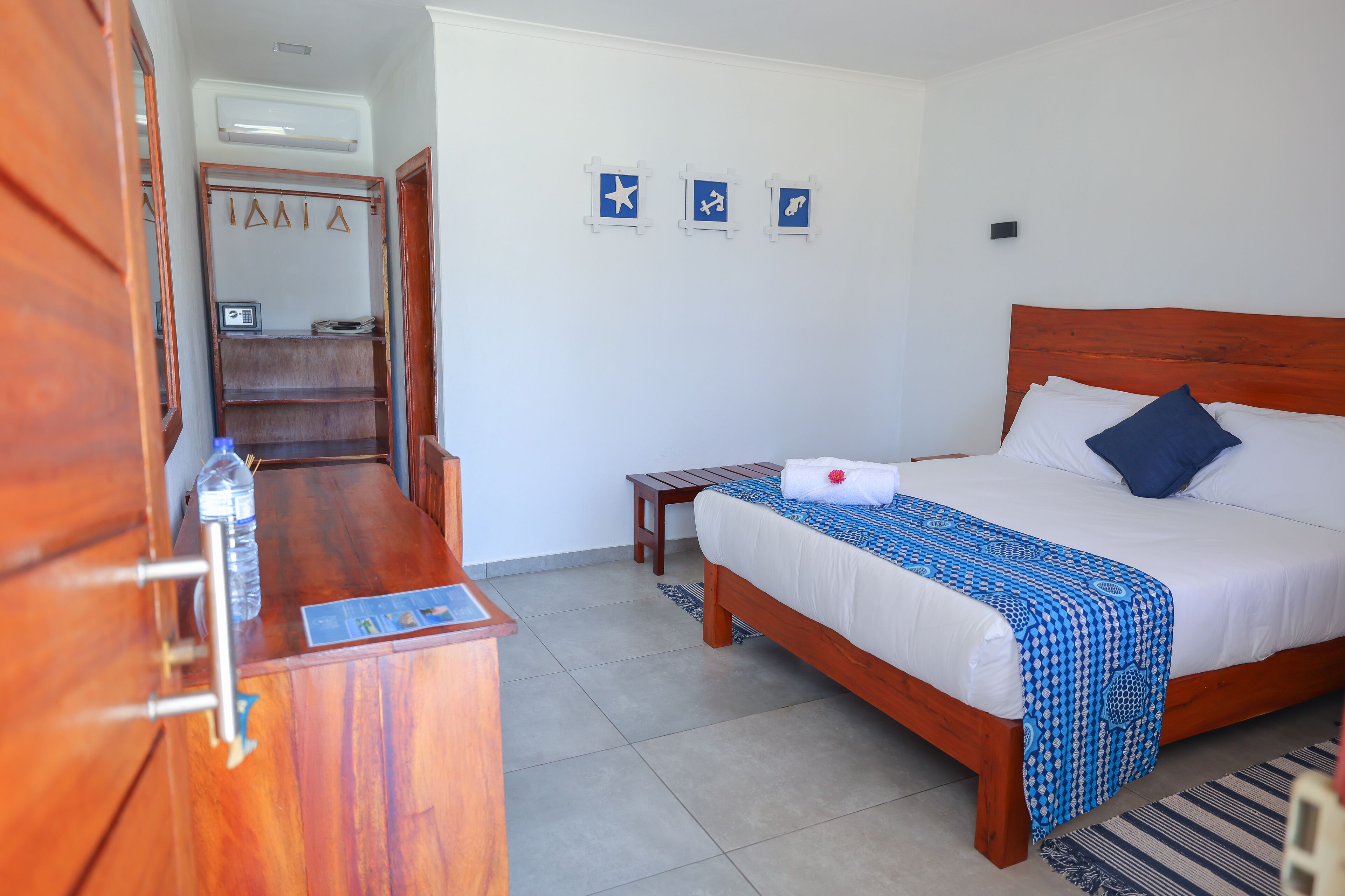 A hotel room with a wooden bed, a wooden dresser, a small bedside table, and a wardrobe. There are three wall-mounted signs above the bed.