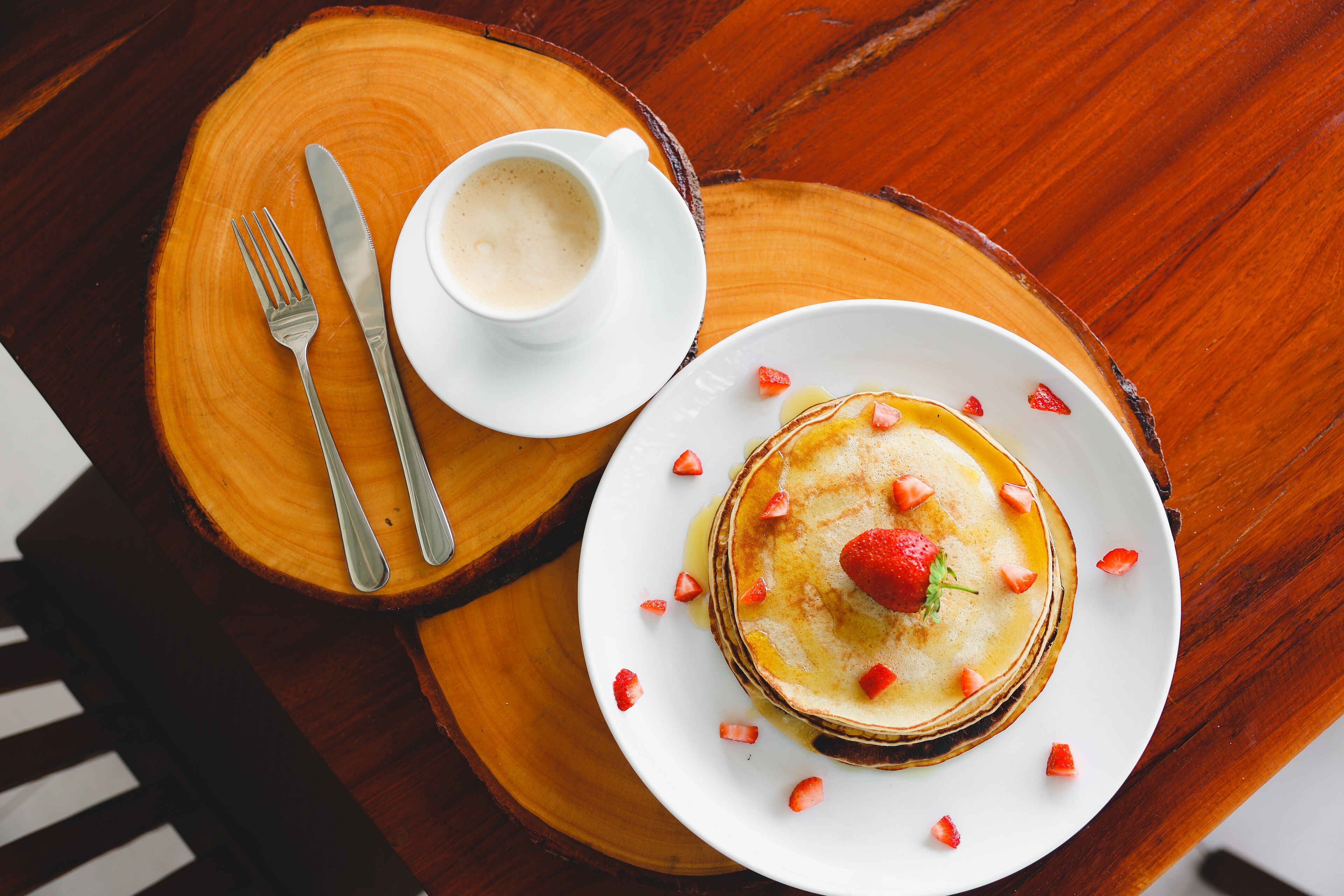 A plate of pancakes with strawberries and syrup, a cup of coffee, and cutlery on a wooden table.