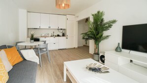 Ein modernes Wohnzimmer mit grauem Sofa, einem weißen Couchtisch und einem Flachbildfernseher. Im Hintergrund ist eine Küche mit weißen Schränken und eine Essecke mit Tisch und Stühlen zu sehen.
