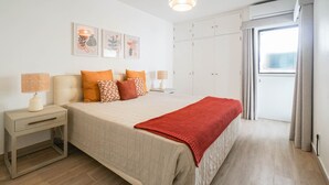 Ein Schlafzimmer mit einem Bett, Nachttischen, einer roten Tagesdecke und einem Fenster mit Vorhängen.