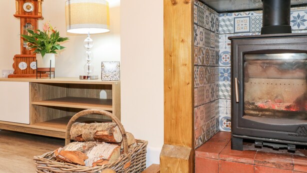 A cozy room with a wooden cabinet, a lamp, a clock, a basket of bread, and a wood-burning stove. - 8 Church Lane (Stoke-on-Trent)