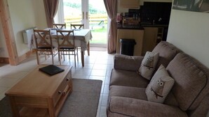 Ein Wohnzimmer mit Couchtisch, braunem Sofa mit Eulen-Kissen, Esstisch mit Stühlen und einer Küchenzeile inklusive Schränke und Spüle.